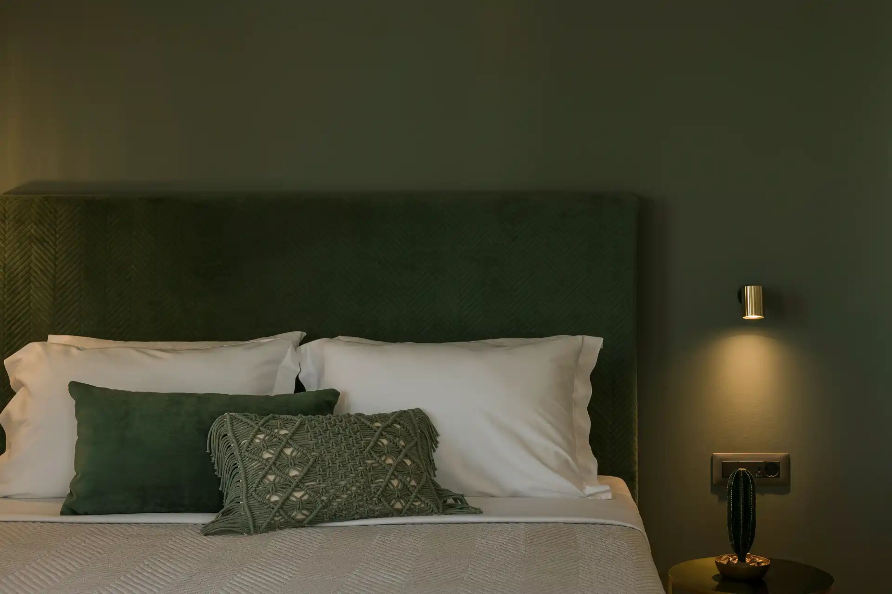 Una cama bien hecha con almohadas blancas y verdes, un plaid de macramé verde y una lámpara fijada a la pared.