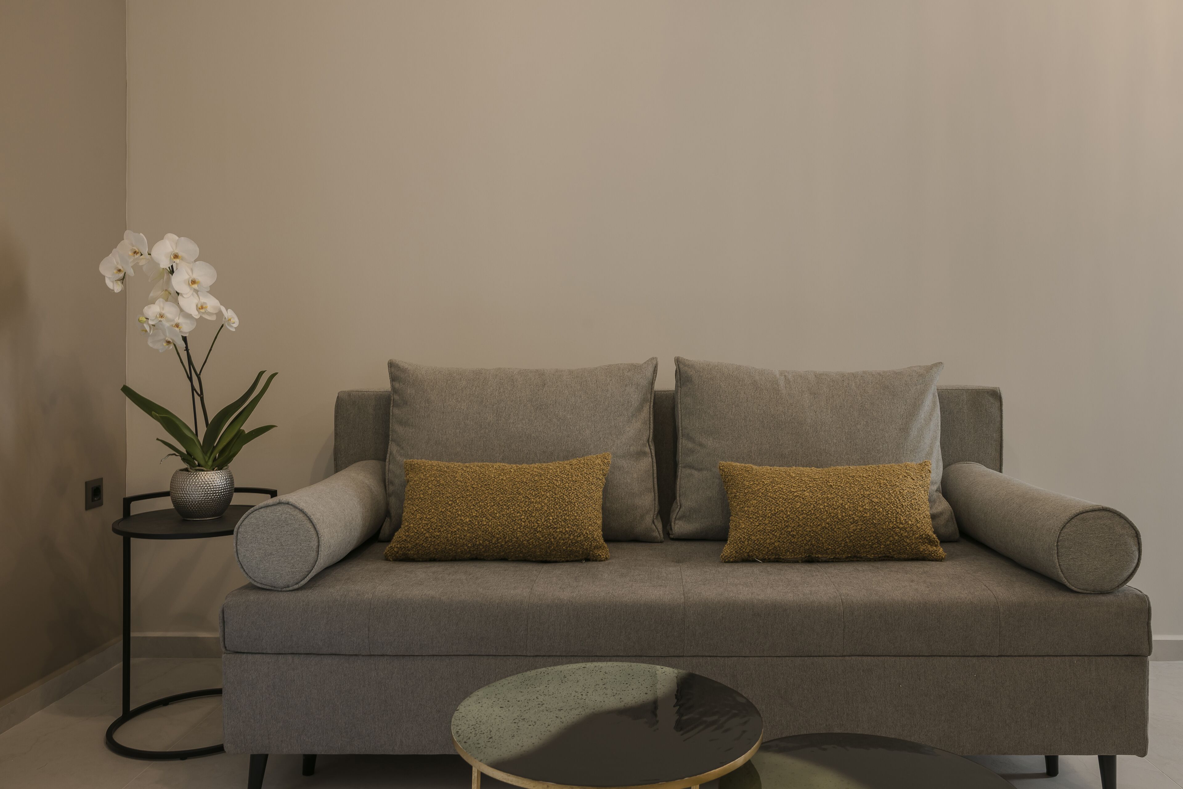 A modern living room with a grey sofa, two mustard-colored cushions, a small round table, and a potted plant with white flowers.