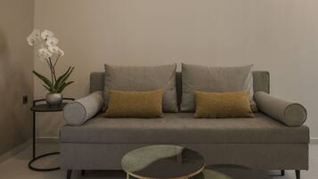 A modern living room with a grey sofa, two mustard-colored cushions, a small round table, and a potted plant with white flowers.
