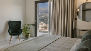 Une chambre d’hôtel avec un lit, un fauteuil vert, une plante en pot, une vue sur les montagnes par la porte d’un balcon et un miroir rond accroché au mur.