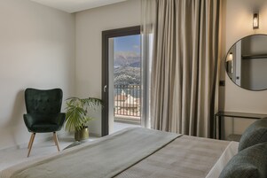 Une chambre d’hôtel avec un lit, un fauteuil vert, une plante en pot, une vue sur les montagnes par la porte d’un balcon et un miroir rond accroché au mur.