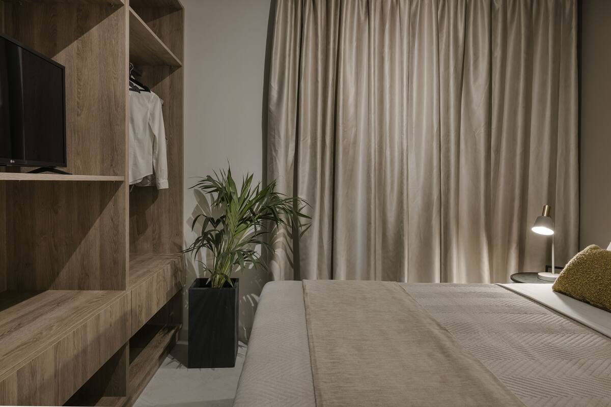 Um quarto moderno com um suporte de madeira para TV, uma planta em vaso, uma cama com manta bege, um abajur e uma janela grande com cortinas.