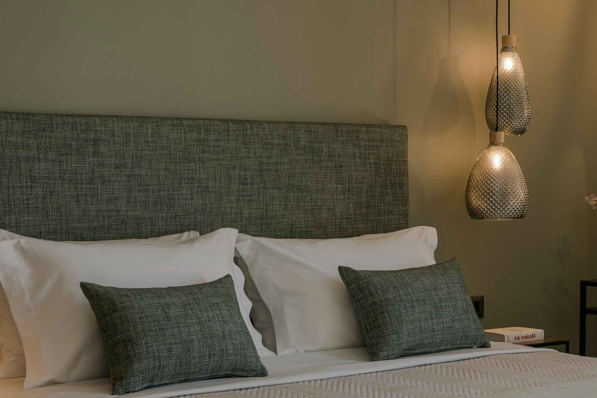 Cama bem arrumada com lençóis brancos e duas almofadas verdes, cabeceira verde texturizada e duas luminárias pendentes.