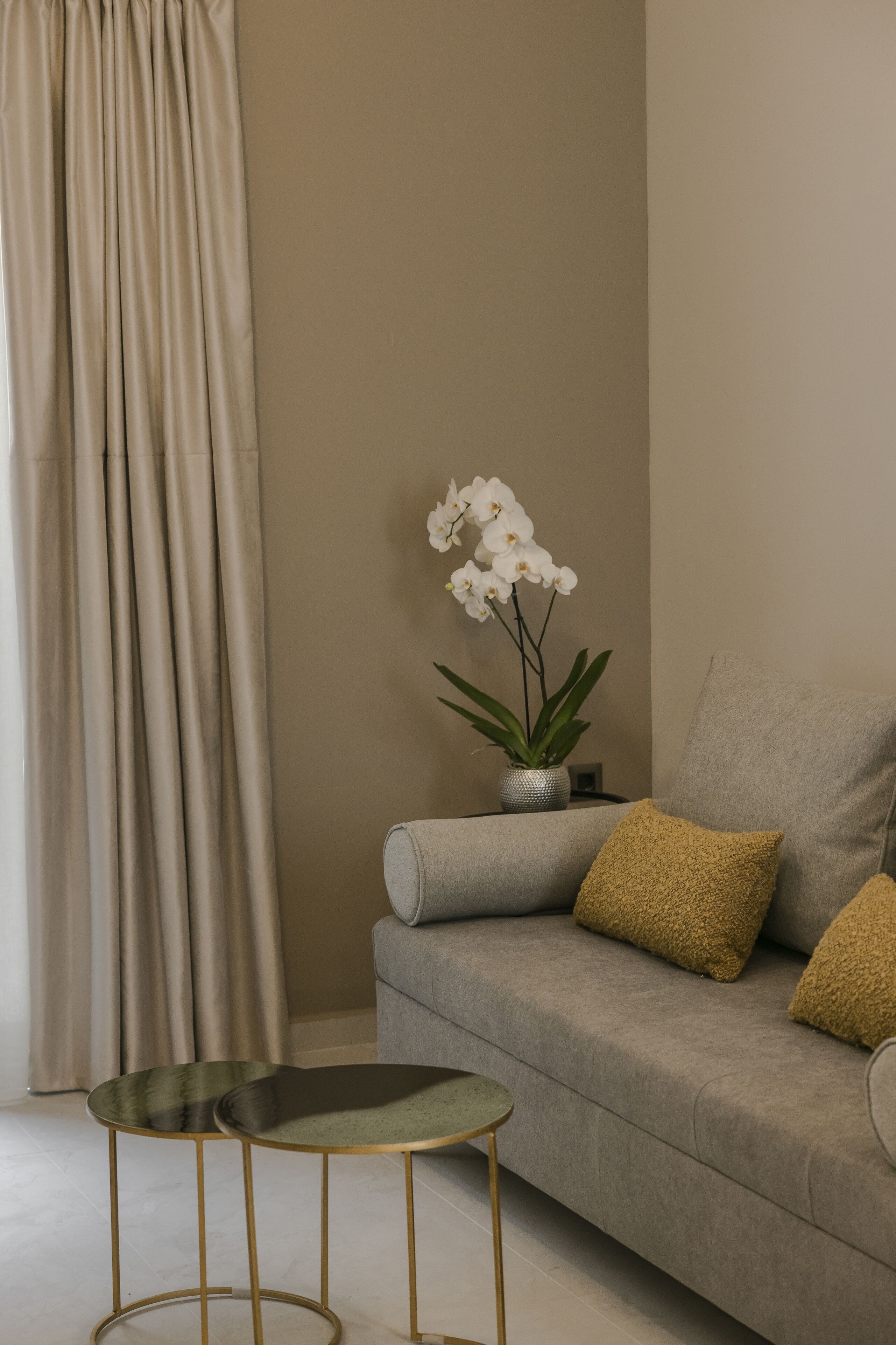 Un salón con un sofá gris, dos mesas redondas con patas doradas, una orquídea en maceta y una cortina color beige.