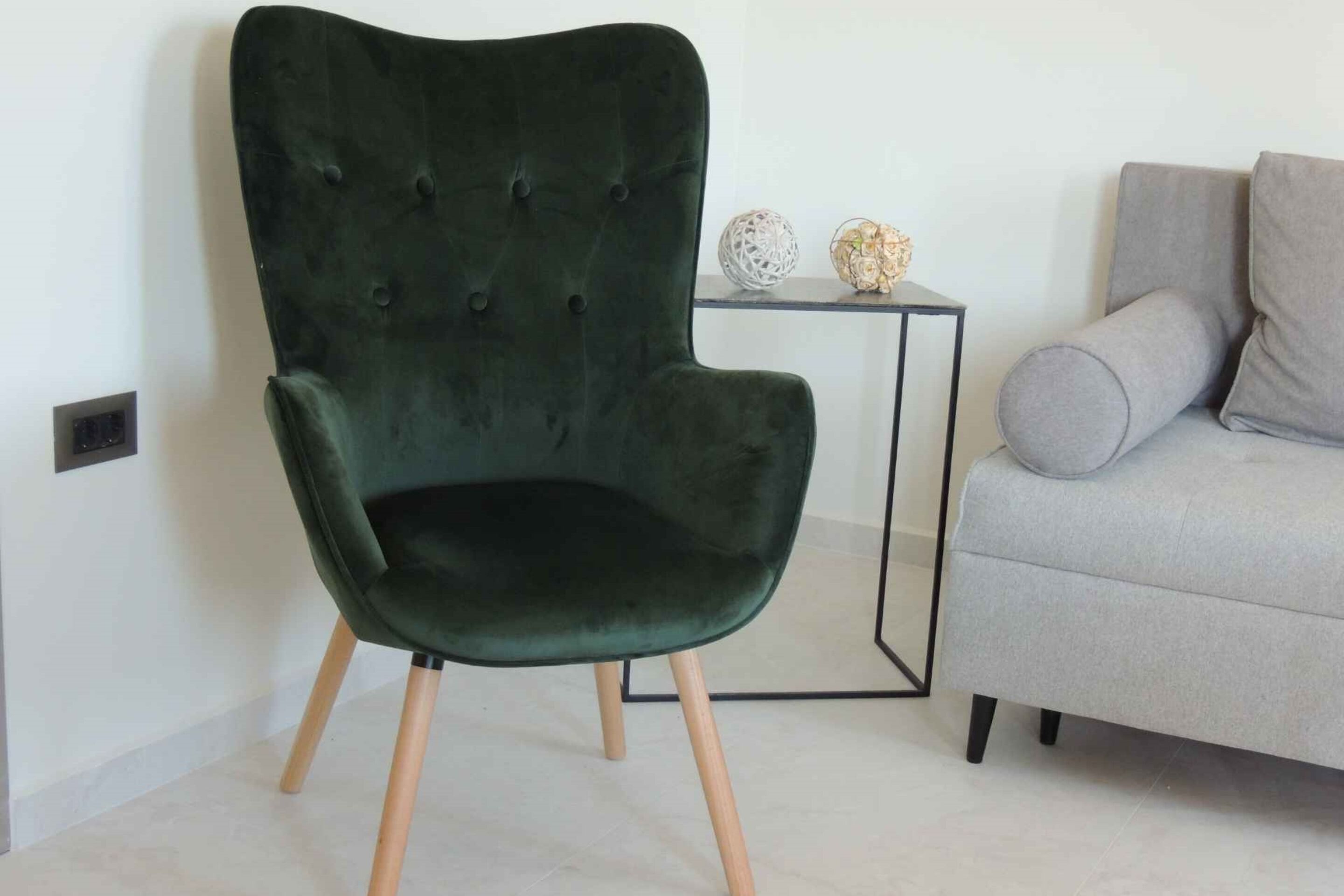 A dark green upholstered chair with wooden legs, positioned next to a grey sofa and a small black side table with decorative items.