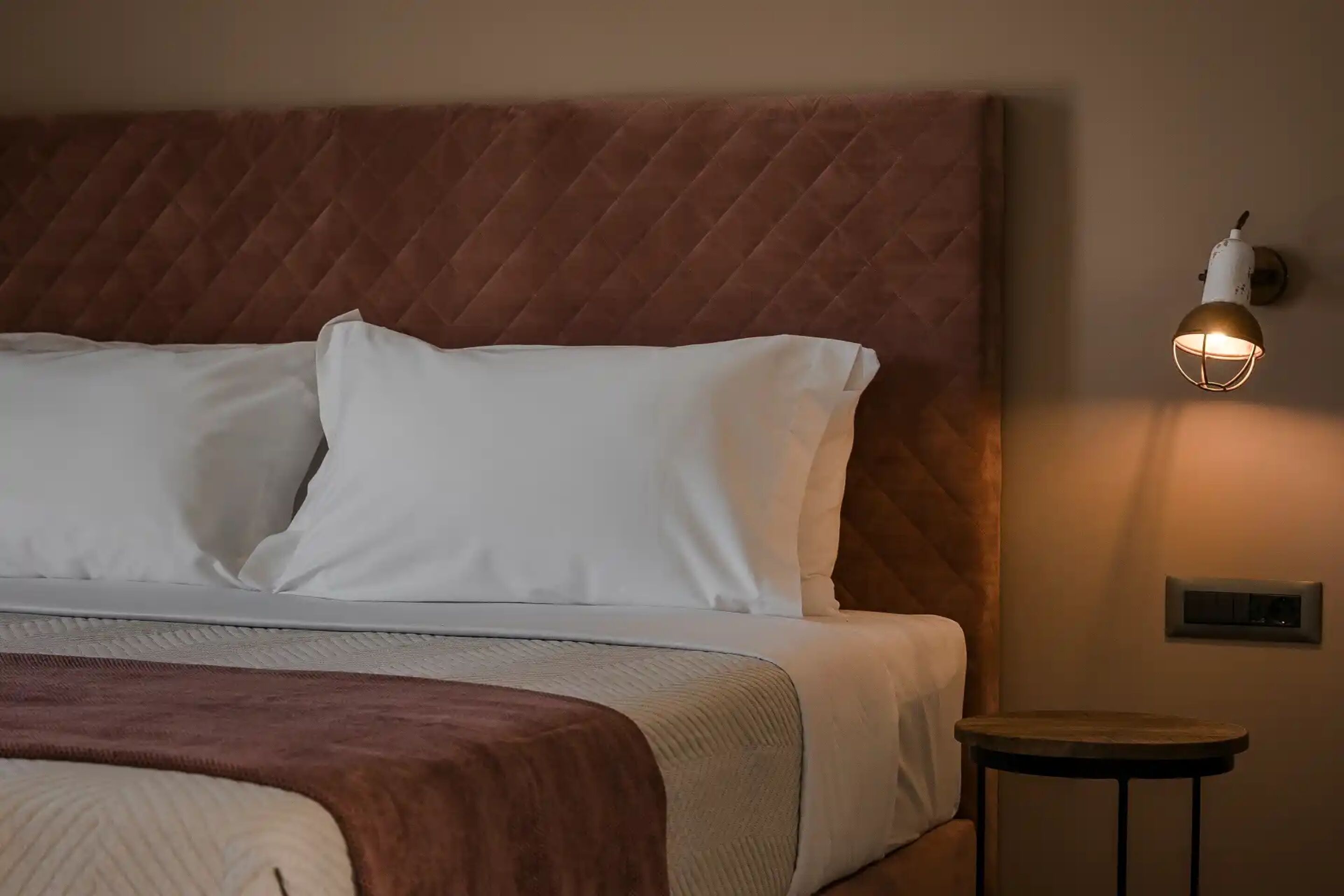 Un lit bien fait, avec du linge de lit blanc et une tête de lit marron. Une petite table ronde se trouve à côté du lit, et un luminaire est fixé au mur.