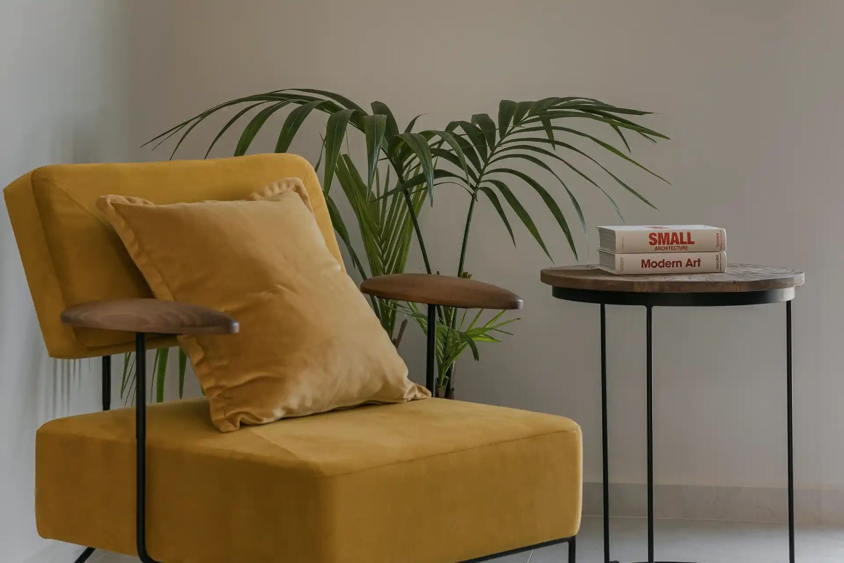 A mustard-colored armchair with a wooden armrest and a matching cushion, positioned next to a small round table with two books titled 'Small Architecture' and 'Modern Art'. A large green plant is in the background.