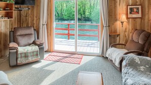 Una camera accogliente con soffitto spiovente, pareti in legno, ventilatore a soffitto, TV a schermo piatto, divano, poltrona e vista su un fiume attraverso una porta in vetro.