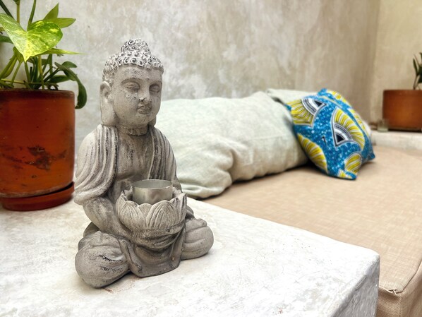 A statue of a seated figure holding a bowl, placed on a textured surface with a potted plant to the left.