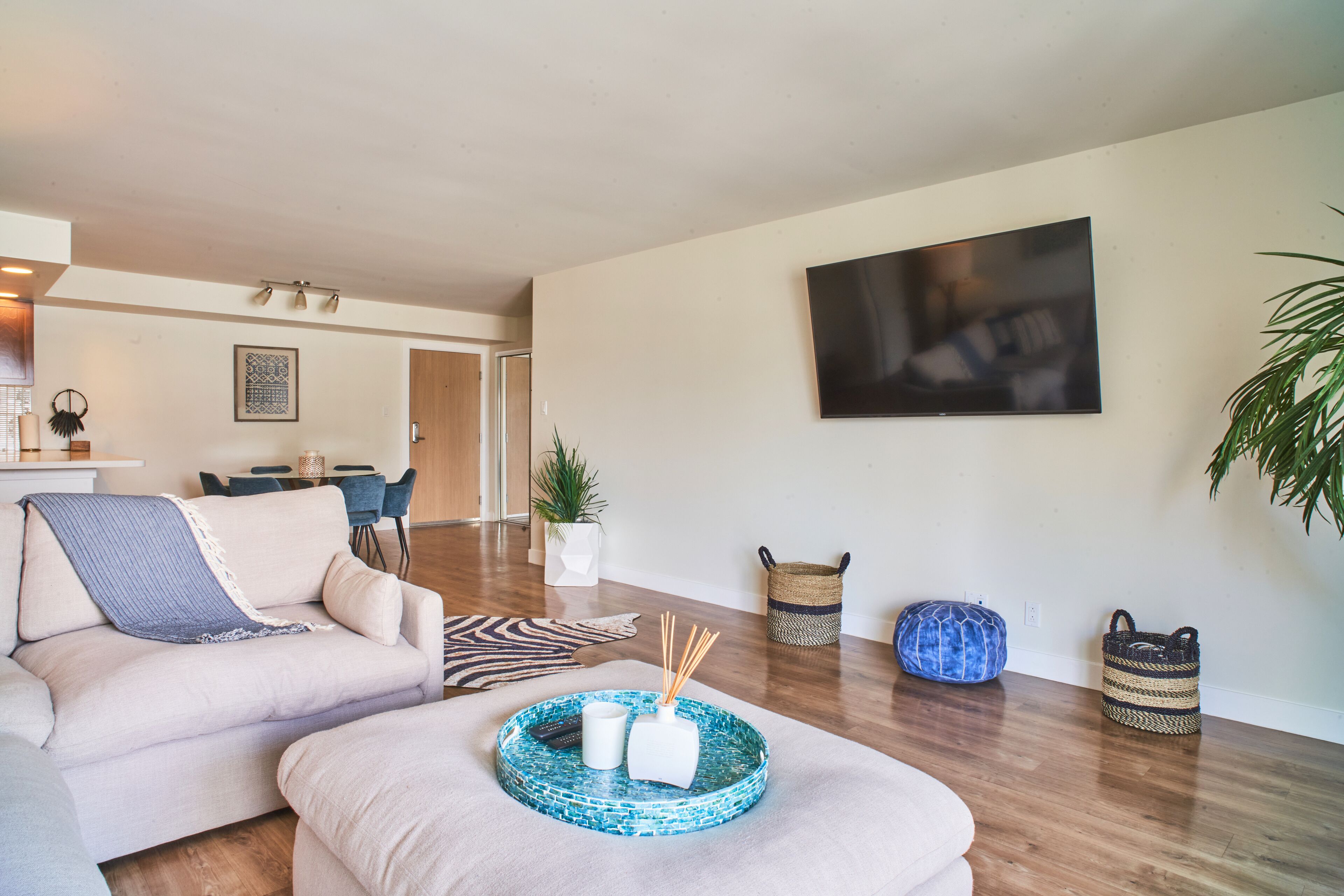 A modern living room with a sofa, ottoman, and a coffee table with decorative items. There is a dining area in the background with a table and chairs.