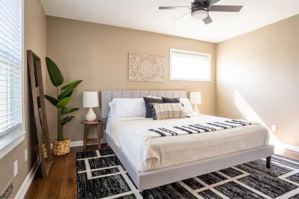 Quarto com cama, ventoinha no teto, ar-condicionado fixado na parede, janela com persianas e planta em vaso.