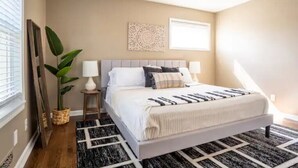 A bedroom with a bed, a ceiling fan, a wall-mounted air conditioner, a window with blinds, and a potted plant.