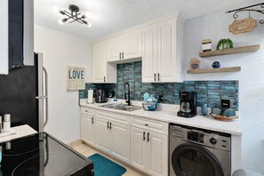 A compact kitchen with white cabinets, a coffee machine, a washing machine, and a blue tiled backsplash.