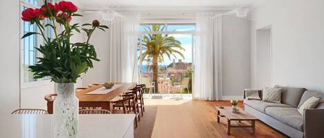 A modern living room with a dining area, a sofa, a coffee table, and a view of a palm tree through large windows.