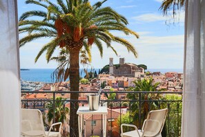 A balcony with two wicker chairs, a small table, and a view of a coastal town with buildings and a clock tower. - Luxury Apartment with Exceptional View (Cannes)