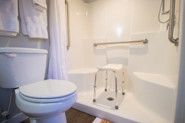 Un baño con inodoro, ducha con barra de apoyo y un taburete pequeño.