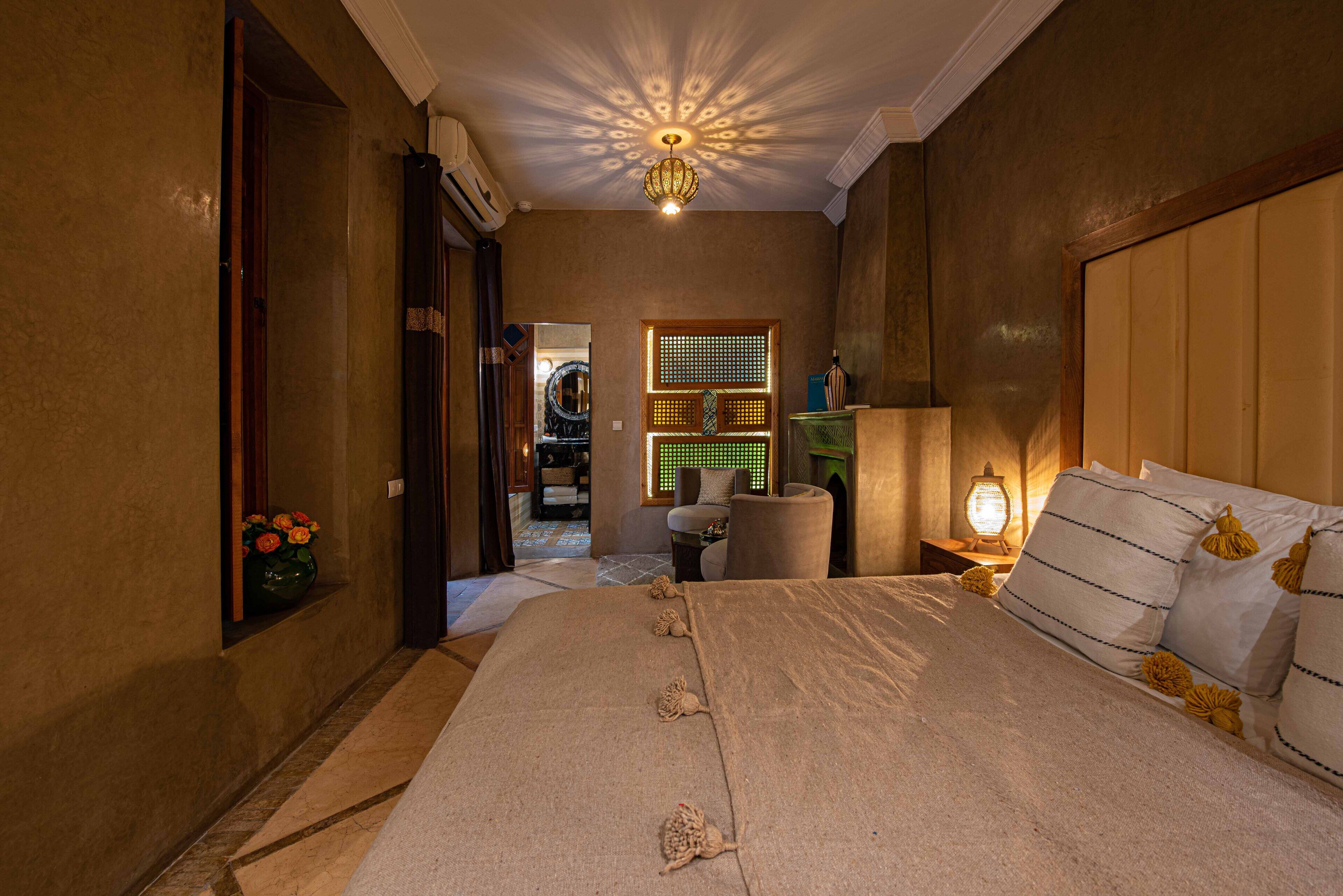 A bedroom with a large bed, a nightstand with a lamp, a chair, and a decorative wall panel.