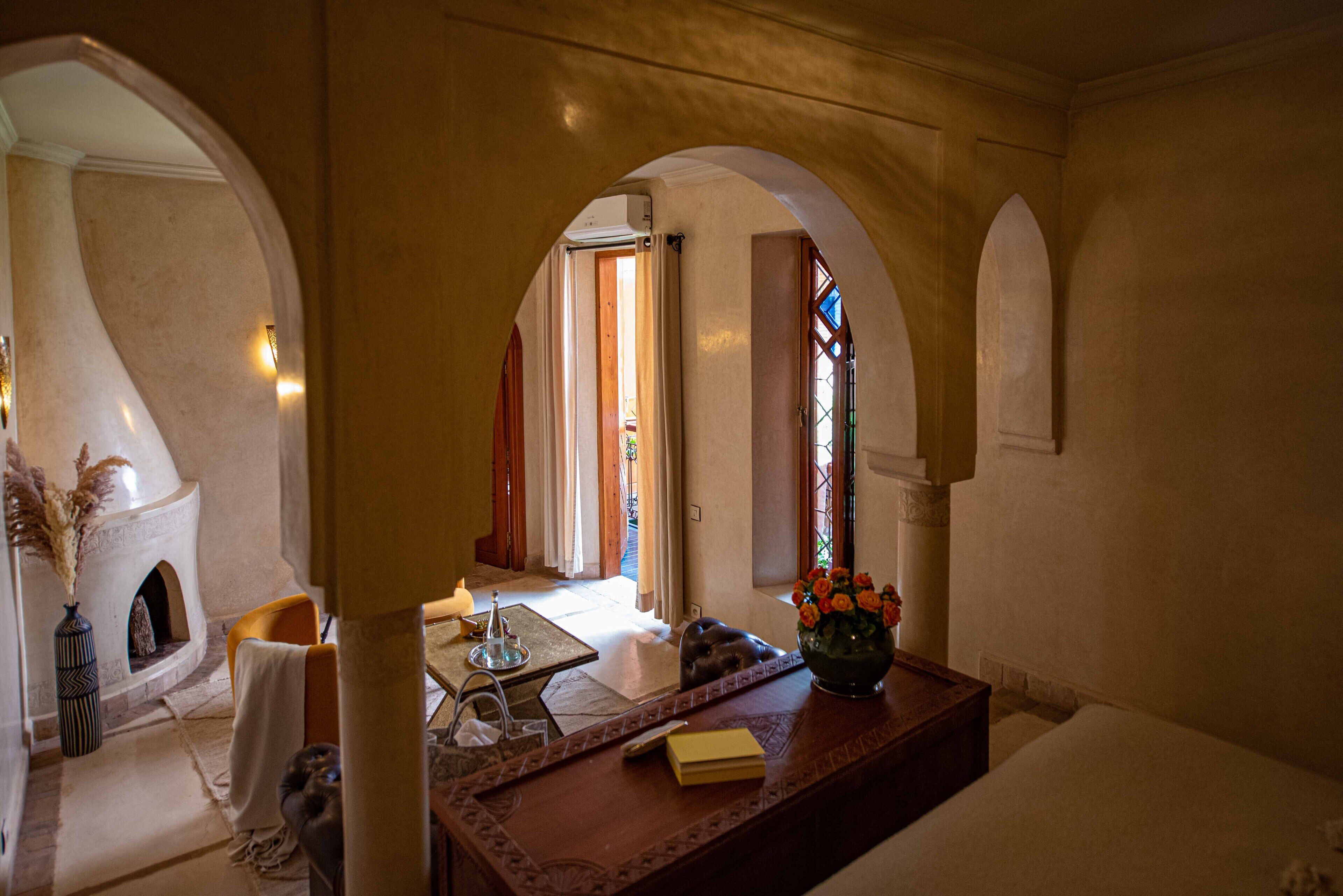 A cozy room with a fireplace, a wooden table with a vase of flowers, and a view through an arched doorway.