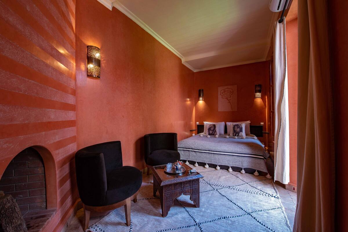 A hotel room with a brick fireplace, a bed with a patterned bedspread, two black armchairs, a small wooden coffee table, and a wall-mounted light fixture.