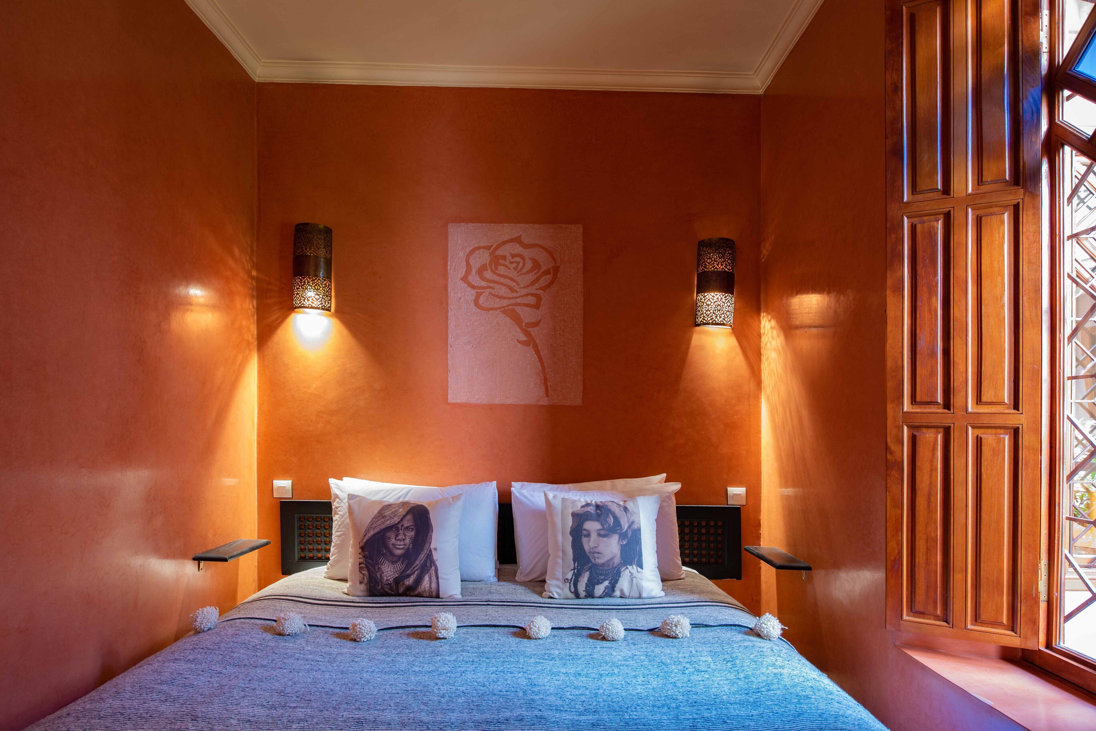 A bedroom with a bed, two wall-mounted lamps, a decorative rose painting, and a window with a view.