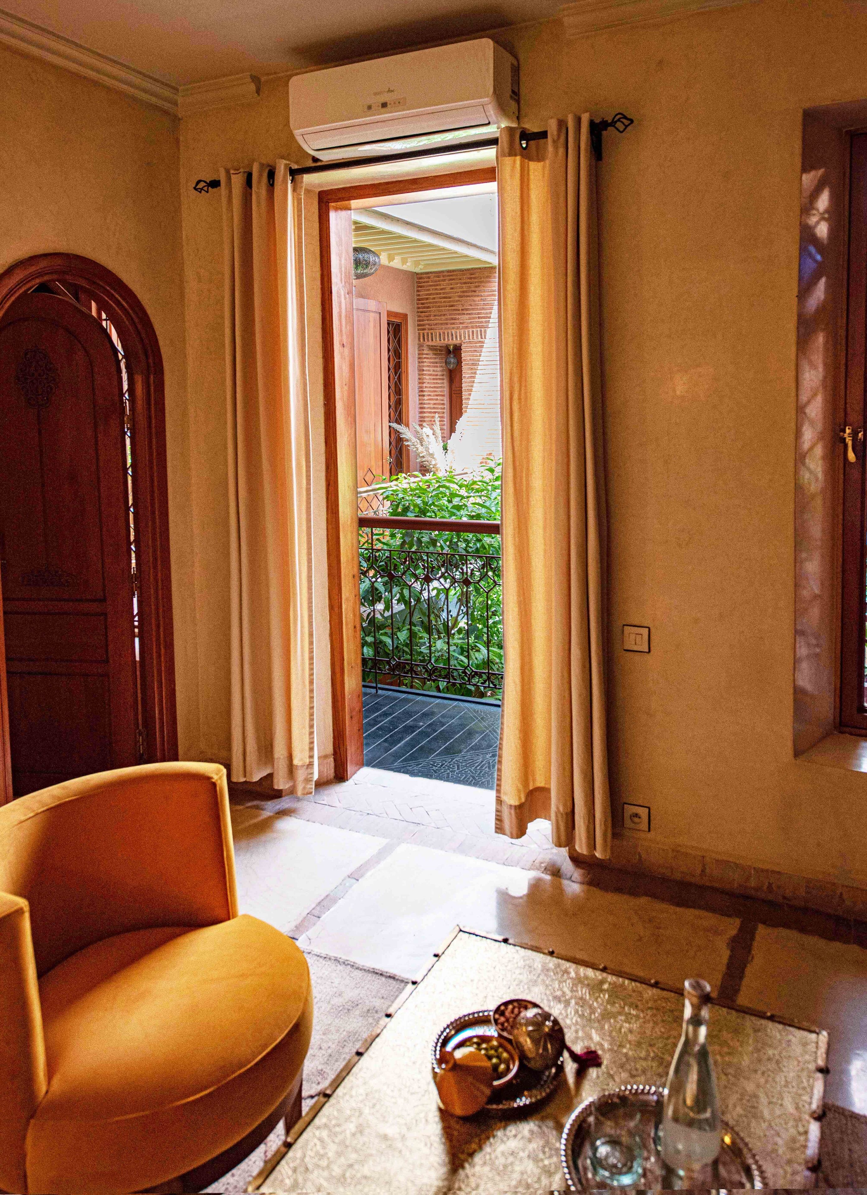 A room with a wooden door, a yellow armchair, a table with a teapot and cups, and a view of a balcony with plants.