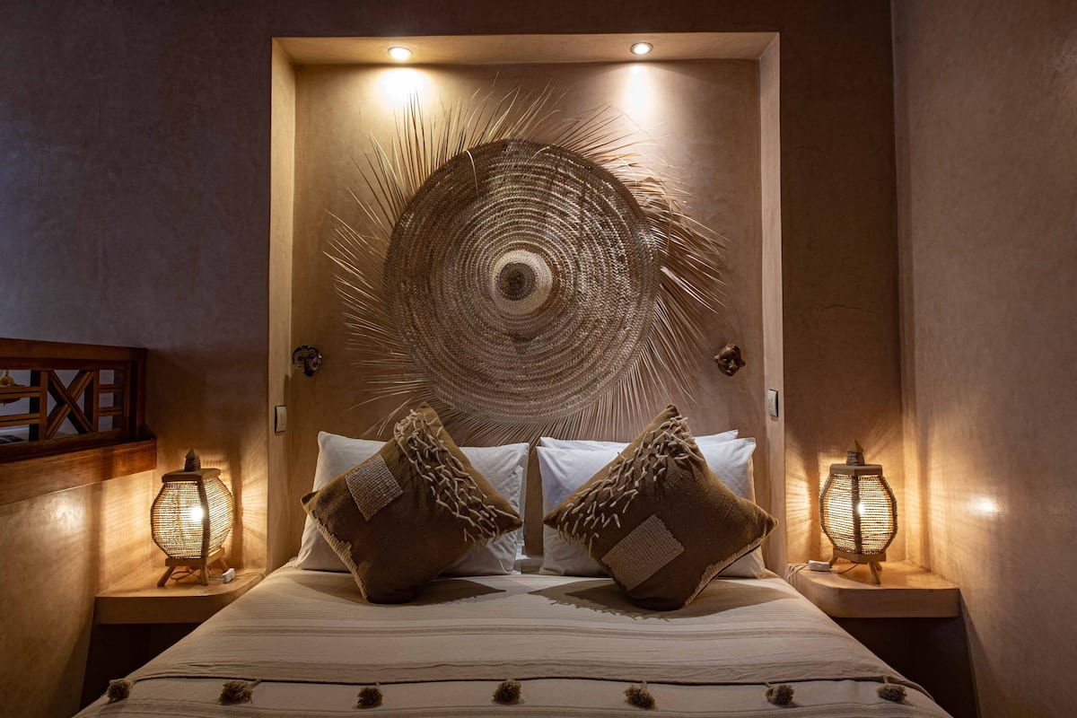 A neatly made bed with two pillows, a wall-mounted circular woven artwork, and two bedside lamps on wooden nightstands.