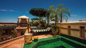 Área de piscina no terraço com piso em azulejo, espreguiçadeiras, espaço de estar coberto e uma palmeira.