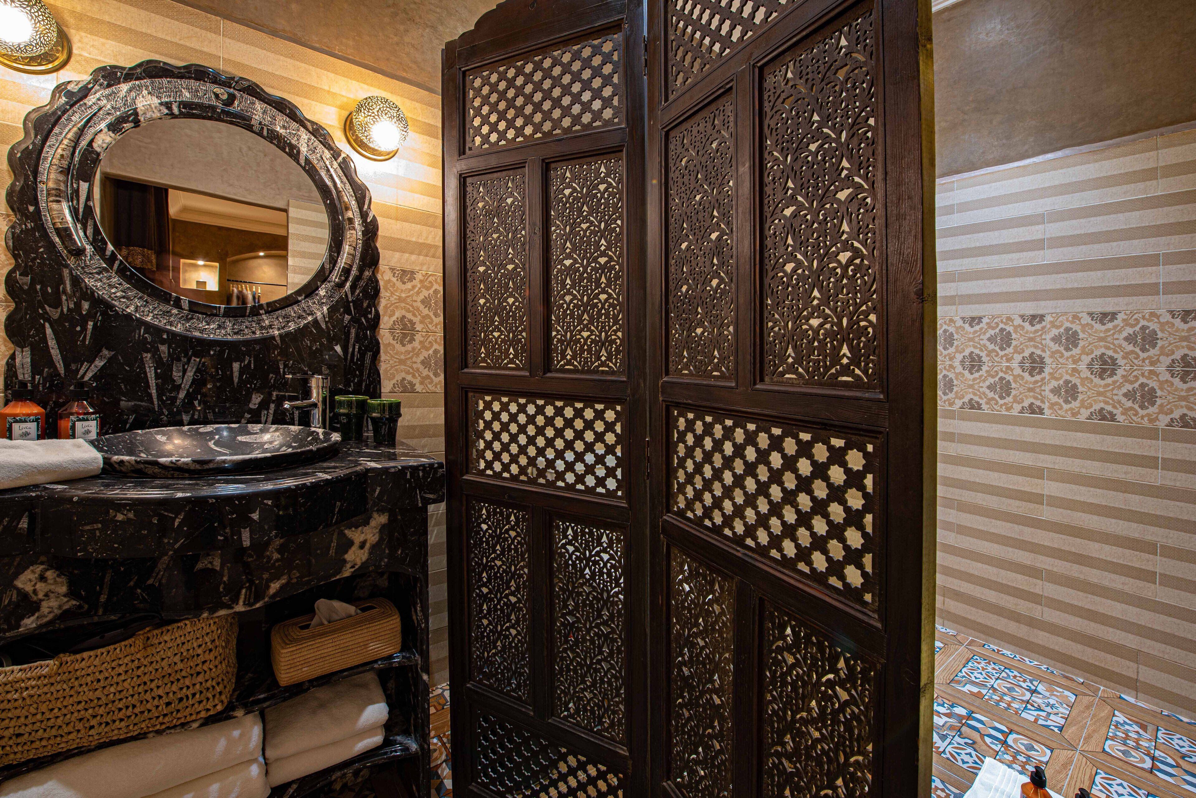 Une salle de bain dotée d’un grand miroir, d’un lavabo avec un plan de toilette en marbre noir et d’une cloison décorative en bois ornée de motifs complexes.