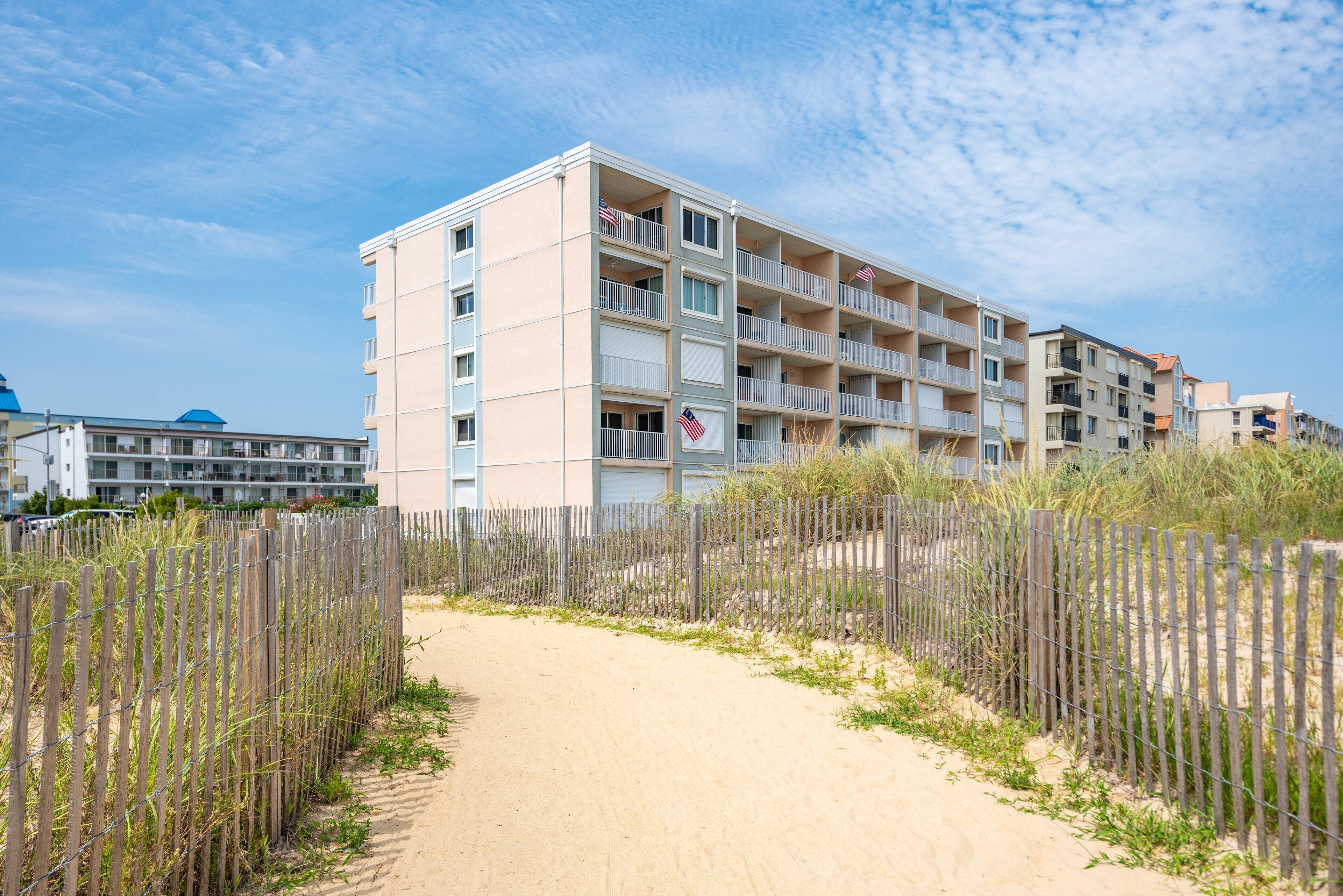 A multi-story hotel with balconies, a sandy pathway, and a wooden fence.