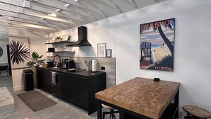 Cocina moderna con una mesa de madera y taburetes de bar, encimera negra y un cuadro grande con una escena de playa.