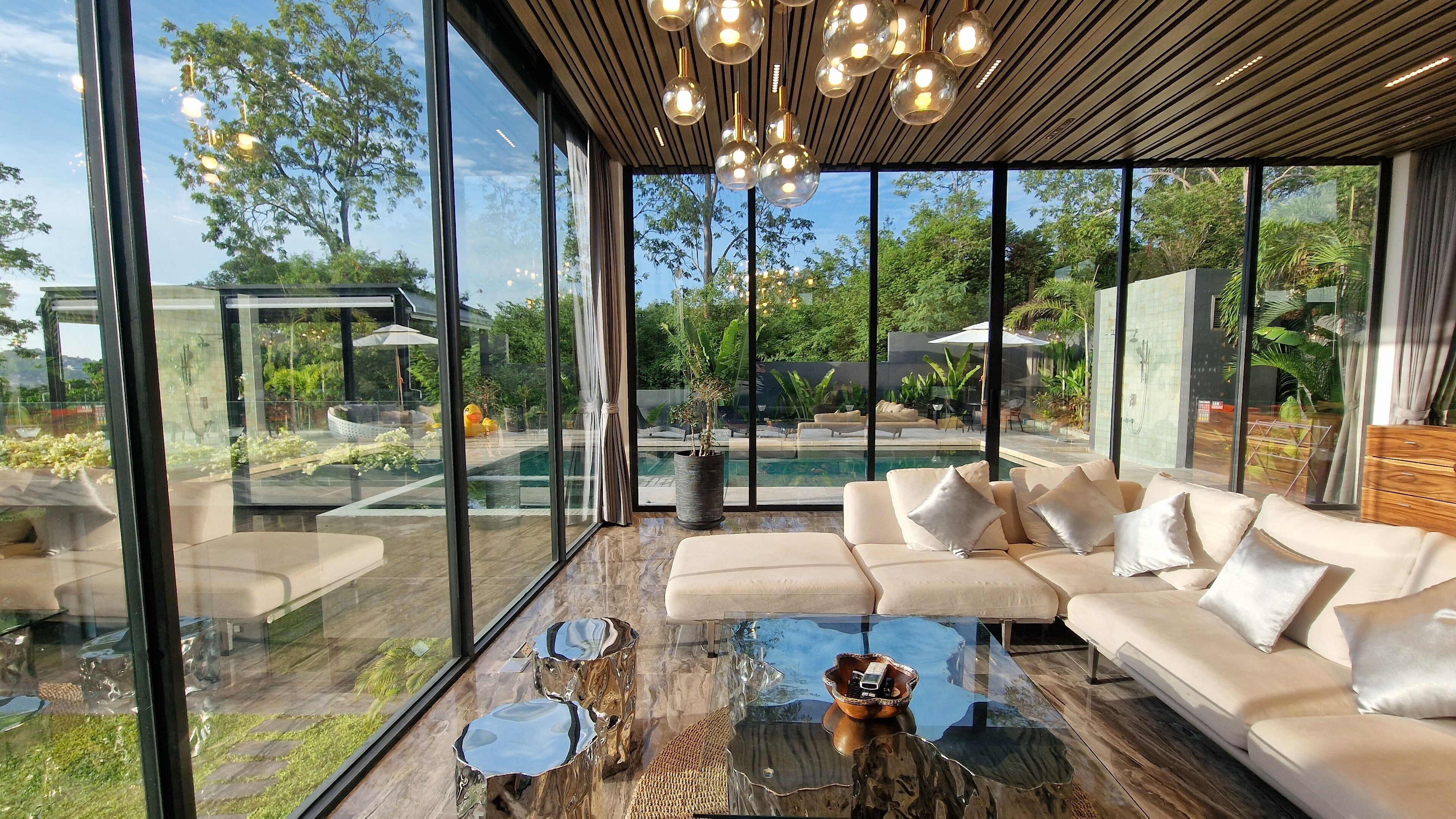A modern living room with a glass coffee table, a beige sofa, and a view of a pool and garden through large windows.