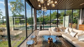A modern living room with a glass coffee table, a beige sofa, and a view of a pool and garden through large windows. - Luxury private 3 Ultra-King BR, with staff, villa sea view 10min walk to beach (Surat Thani)