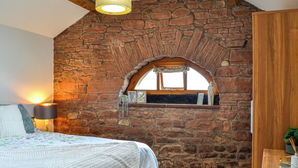 Une chambre avec un mur en pierre, une fenêtre surmontée d’une arche en bois, un lit recouvert d’une couverture blanche, une lampe de chevet et une armoire en bois.