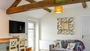 Un salon chaleureux avec une table à manger en bois, un canapé agrémenté de coussins à motifs, un miroir décoratif et un téléviseur fixé au mur.