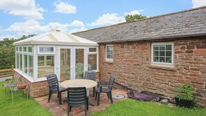 Une véranda avec un toit en verre, un bâtiment en pierre avec un toit en tuiles et une terrasse dotée de mobilier d’extérieur.
