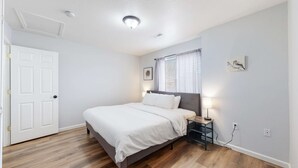 Um quarto com cama, mesa de cabeceira, abajur, janela com persianas, porta e luminária no teto.