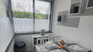 Uma sala de jantar com mesa posta para quatro pessoas, cadeiras brancas, uma janela com persianas e um radiador na parede.
