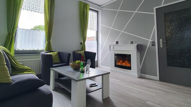Uma sala de estar moderna com lareira, sofá, mesa de centro e vista para o exterior através de grandes janelas com cortinas verdes.