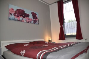 Un dormitorio con cama, mesita de noche, lámpara, ventana con cortinas y un cuadro en la pared.