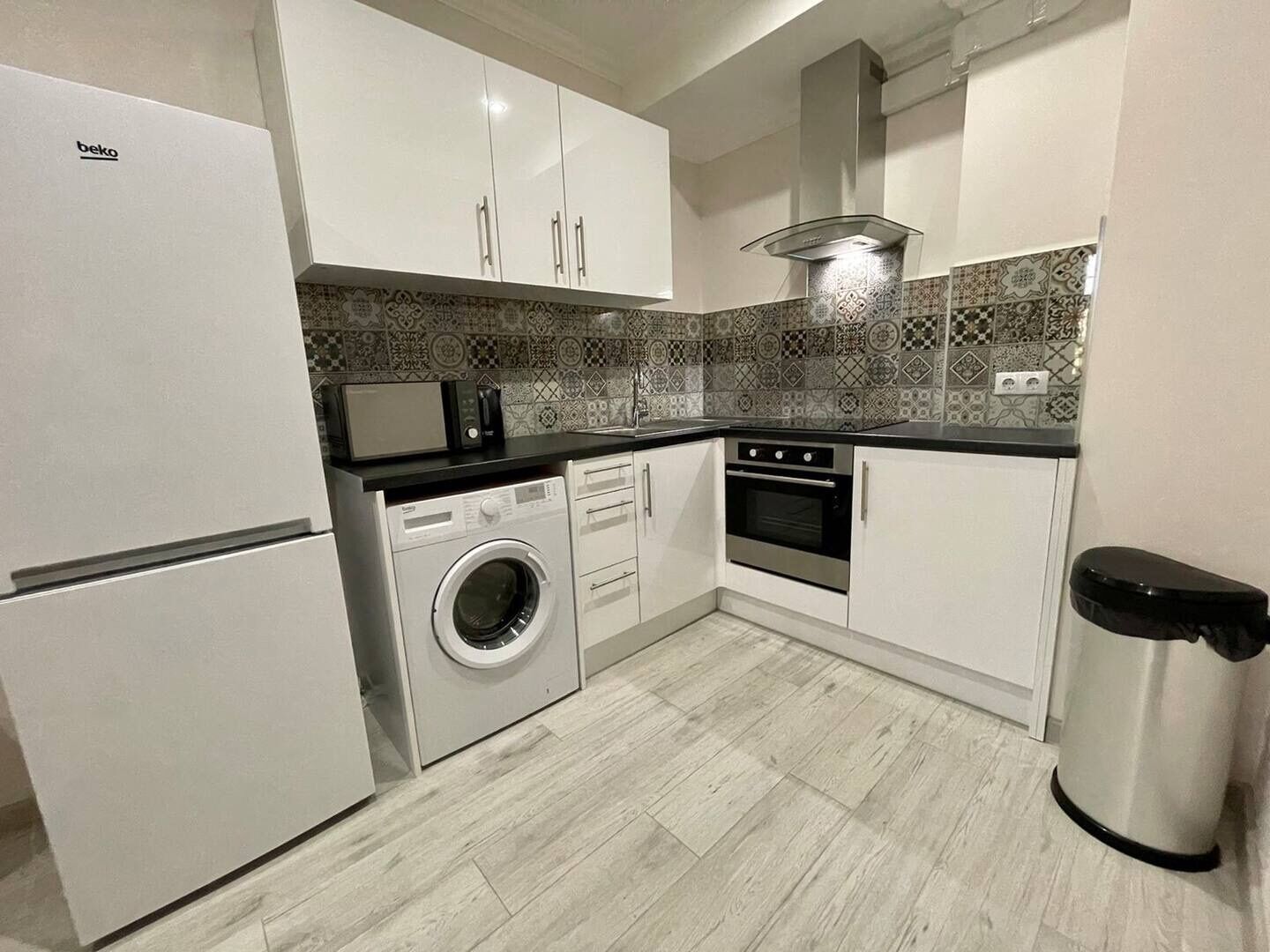 A compact kitchen with a washing machine, oven, microwave, and a trash bin.