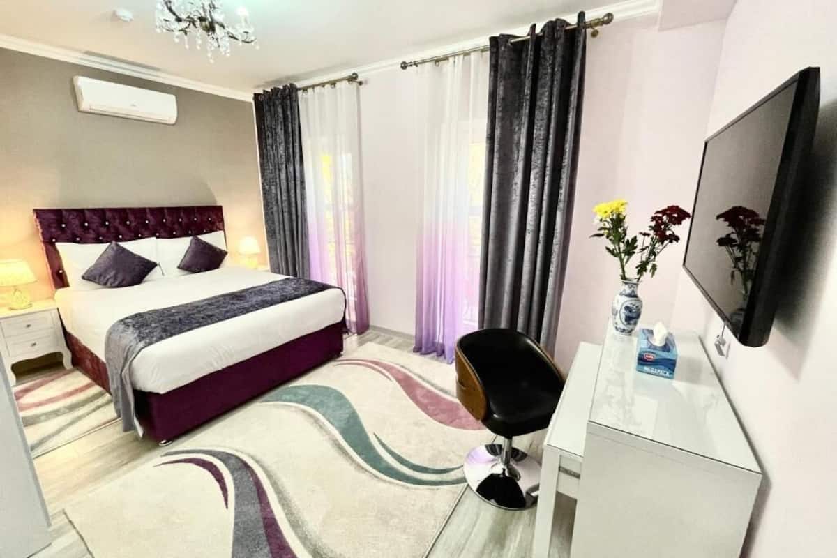Een slaapkamer met een bed, een bureau, een stoel, een televisie en een raam met gordijnen.