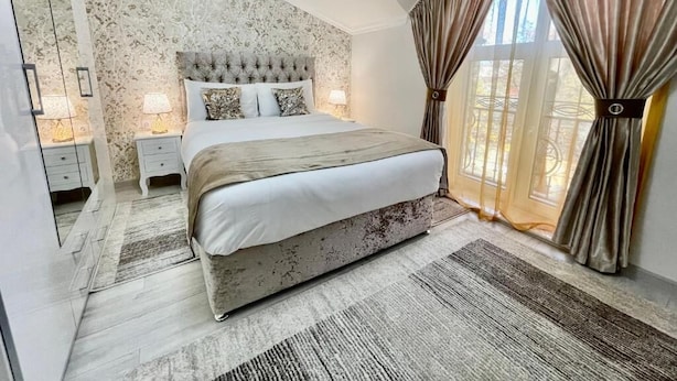 Une chambre à coucher avec une tête de lit capitonnée, un grand lit, des lampes de chevet, une fenêtre avec des rideaux et une vue sur l’extérieur.
