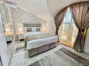 Ein Schlafzimmer mit einer gepolsterten Kopfteil, einem großen Bett, Nachttischlampen, einem Fenster mit Vorhängen und Blick ins Freie.
