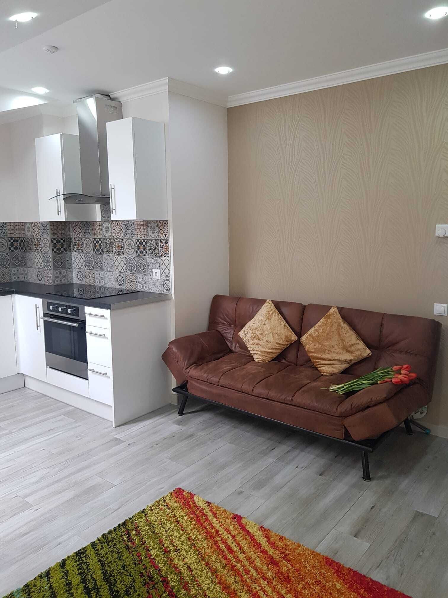 A modern kitchen with white cabinets, a built-in oven, and a patterned backsplash. A brown leather sofa with two cushions is positioned against a textured wall, with a colorful rug on the floor.