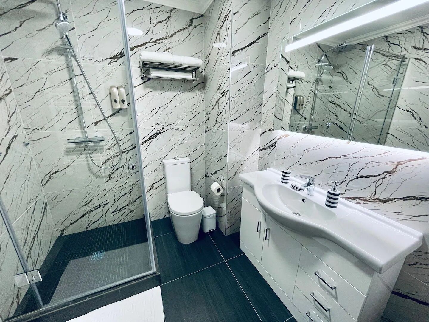 Un baño moderno con ducha envuelta en vidrio, paredes de mármol blanco, un lavamanos blanco y un inodoro.