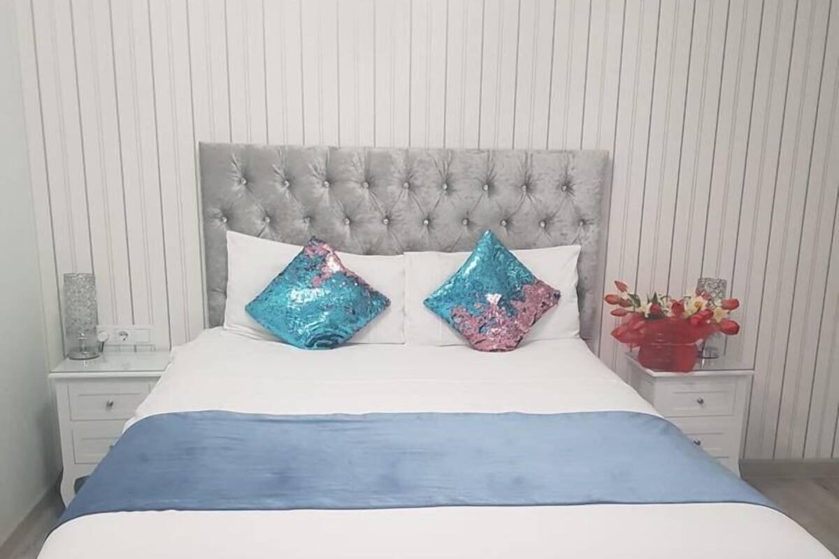 Een bed met een grijs gestoffeerd hoofdeinde, twee decoratieve kussens, een blauwe deken en een nachtkastje met een bloevas.