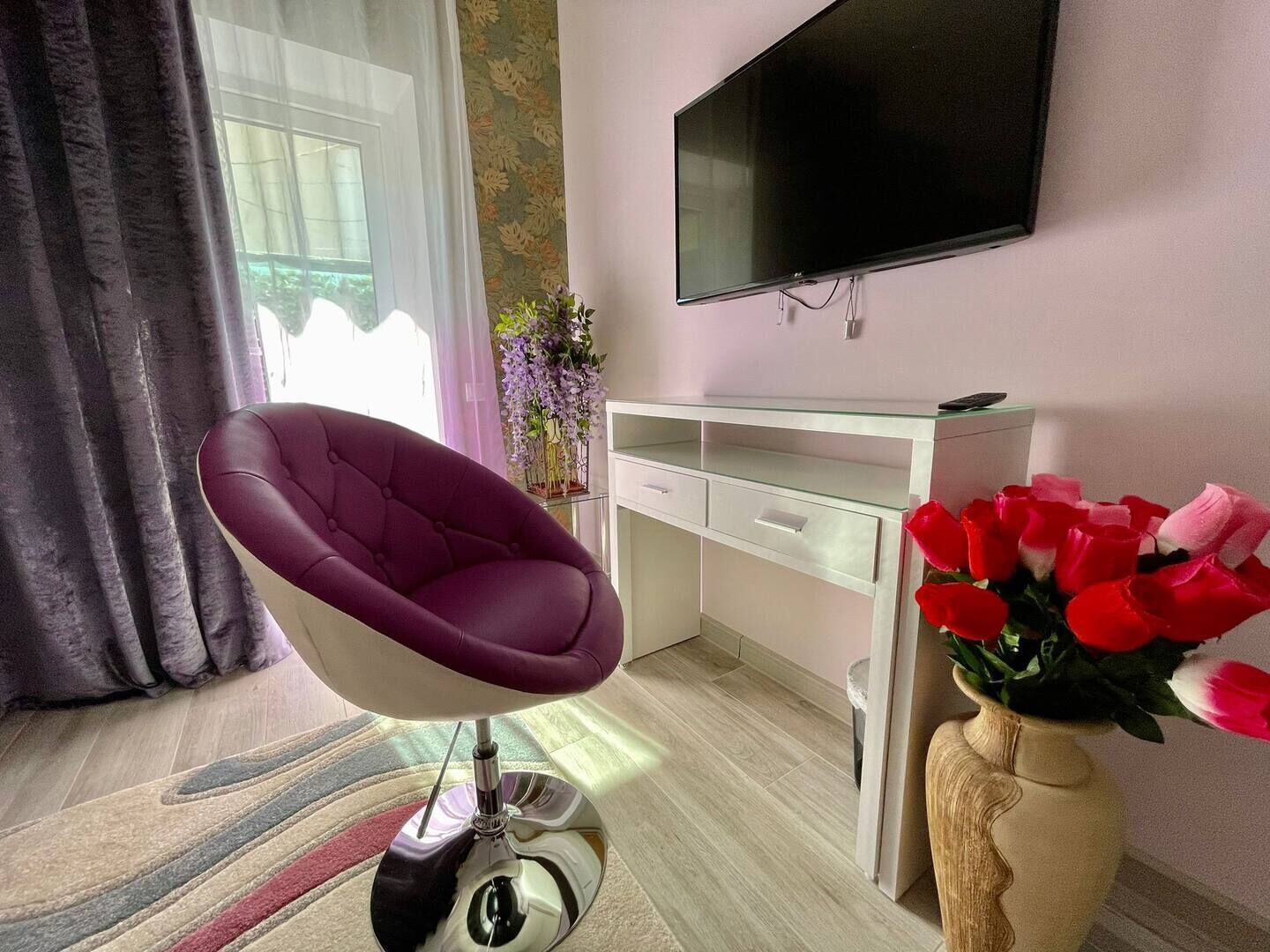 A modern living room with a purple chair, a TV mounted on the wall, a white desk, and a vase with red flowers.