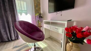 A modern living room with a purple chair, a TV mounted on the wall, a white desk, and a vase with red flowers.