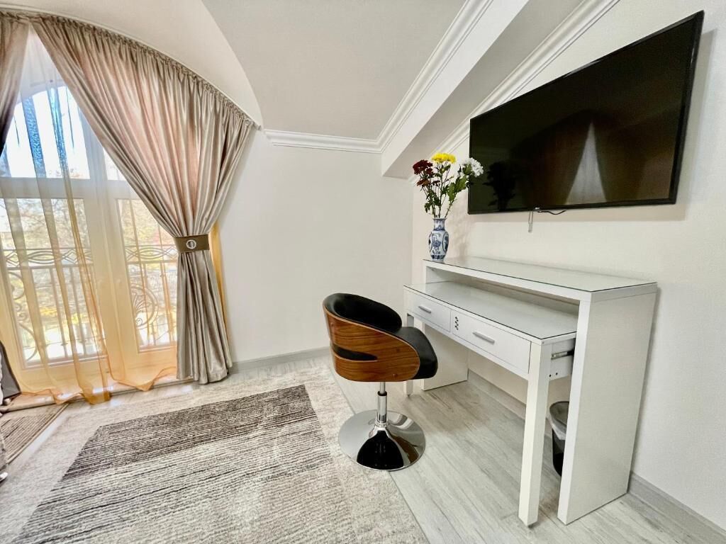 A modern room with a white desk, a black chair, a TV mounted on the wall, and a window with curtains.