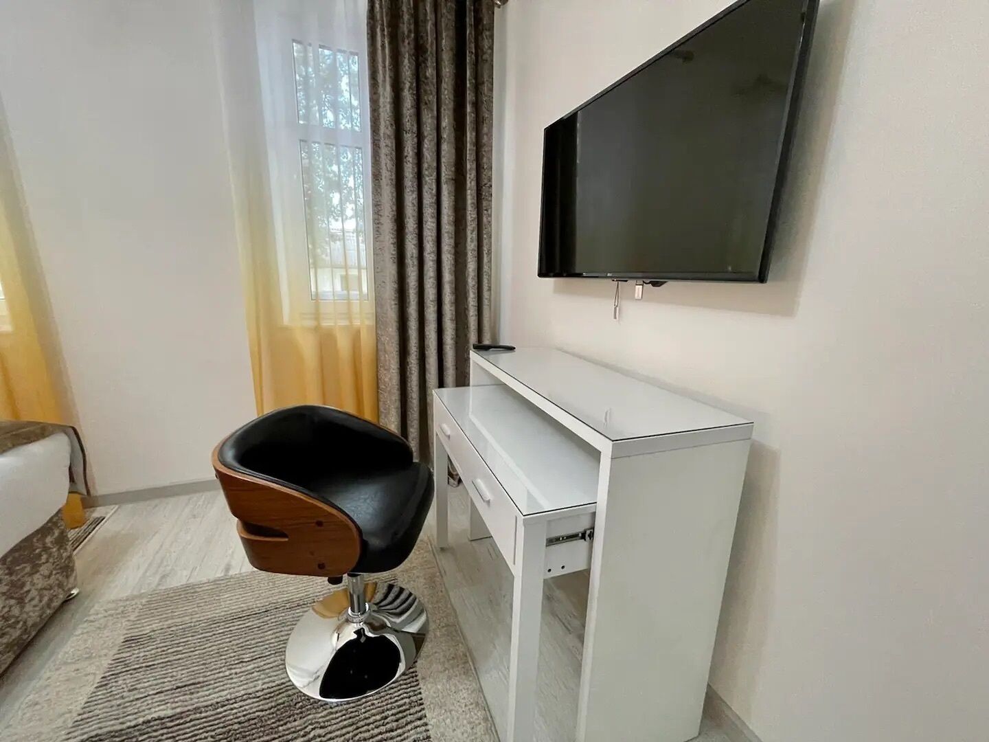 A modern room with a white desk, a black chair, a television mounted on the wall, and a window with curtains.